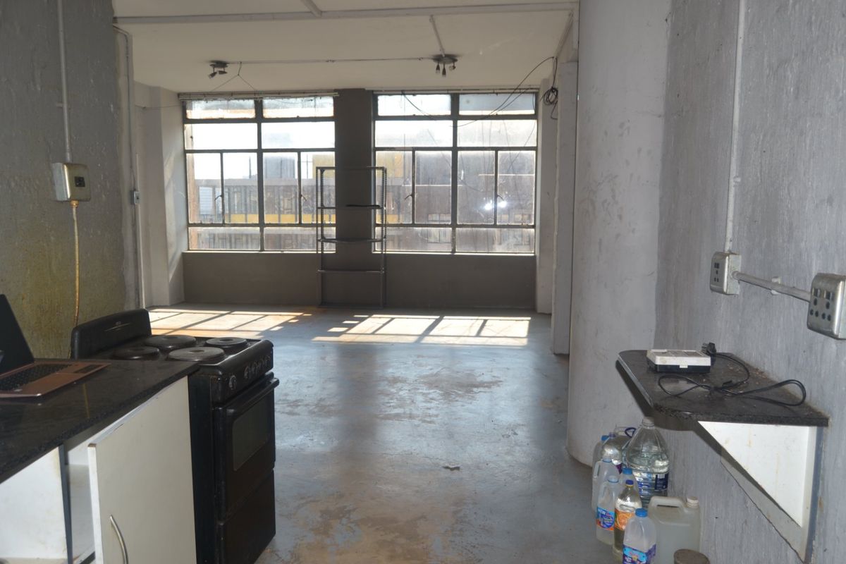 An open-plan living and kitchen area featuring industrial-style windows, exposed concrete walls, and concrete flooring, offering a blank canvas for renovation.