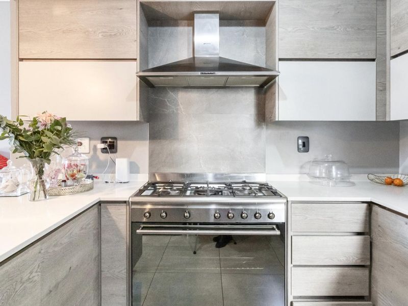 Modern kitchen featuring light wood-grain cabinetry, stone countertops, a stainless steel gas stove, and a matching extractor fan with a sleek tiled backsplash.
