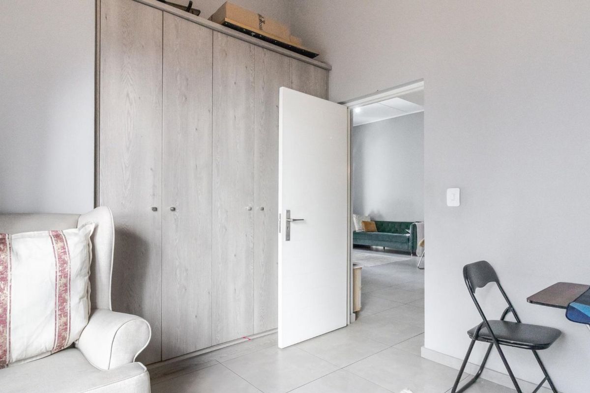 A bedroom featuring light wood-finish built-in cupboards and neutral tiled flooring, with a doorway leading to an adjacent living area.