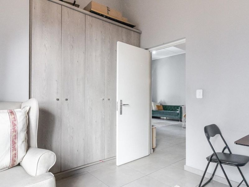 A bedroom featuring light wood-finish built-in cupboards and neutral tiled flooring, with a doorway leading to an adjacent living area.