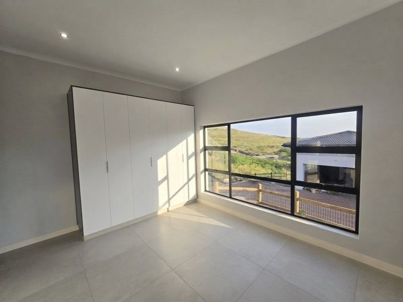 A bright, modern bedroom featuring neutral tiled flooring, built-in cupboards, and large windows offering scenic views.
