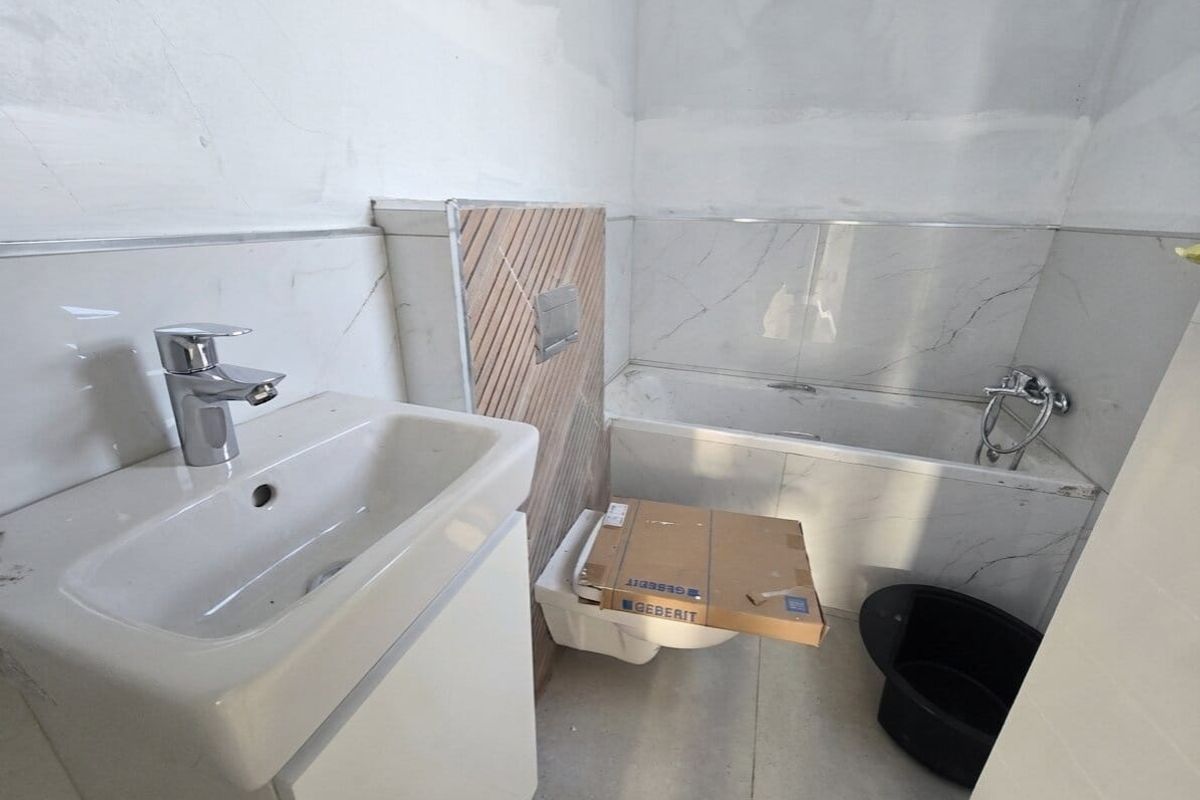 A bathroom under construction featuring a wall-mounted vanity, built-in bathtub, and wall-mounted toilet with marble-effect tiling and unfinished wall surfaces.