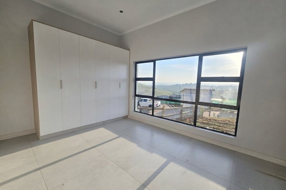 A bright, modern bedroom featuring neutral tiled flooring, ample built-in cupboard space, and a large window offering scenic views.