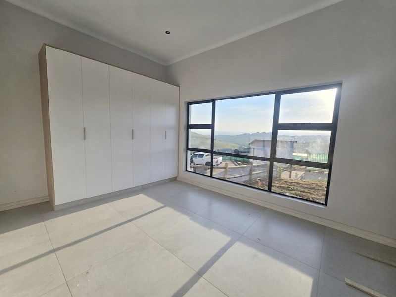 A bright, modern bedroom featuring neutral tiled flooring, ample built-in cupboard space, and a large window offering scenic views.