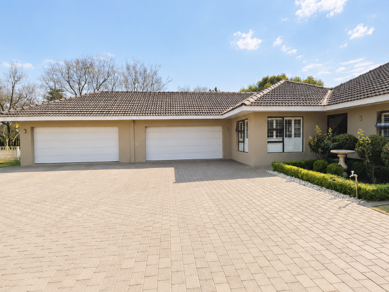 A well-maintained front exterior featuring a spacious paved driveway, double garage, and neat garden landscaping with established shrubs.