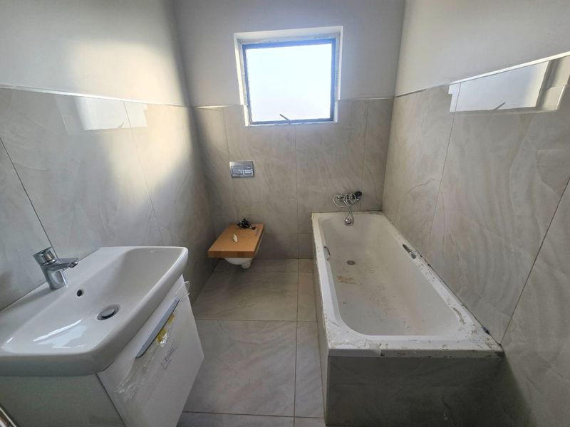 A modern, unfinished bathroom featuring a wall-mounted toilet, bathtub, and vanity basin with neutral wall and floor tiling.