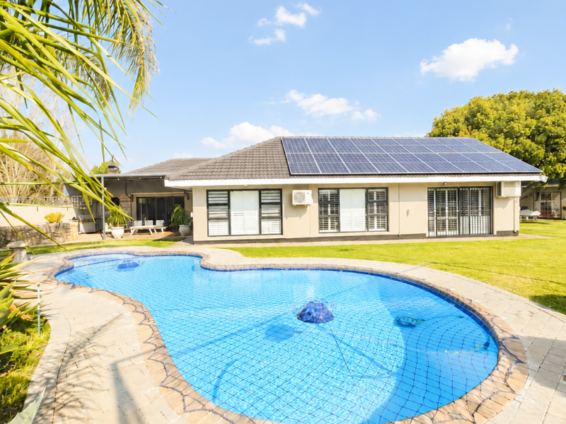 A well-maintained backyard featuring a kidney-shaped swimming pool with a safety net, surrounded by paved decking and a house equipped with solar panels.