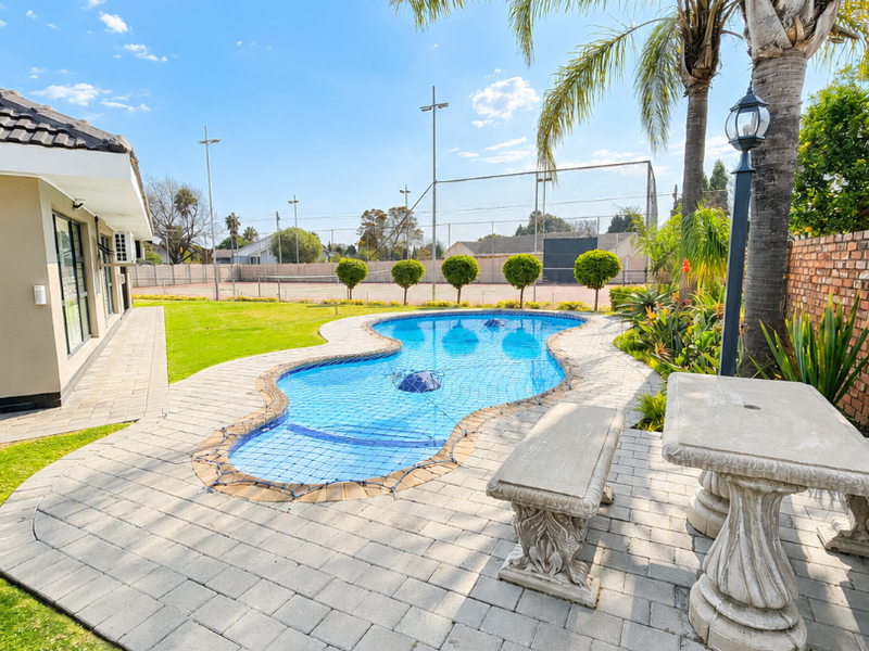 A well-maintained outdoor pool area featuring a paved patio, established palm tree, and views of the adjacent tennis court.