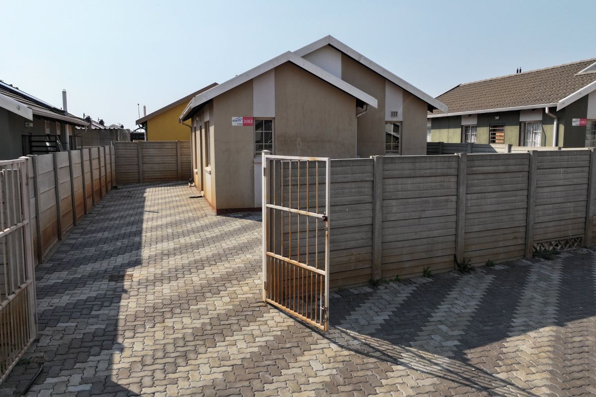 Rear exterior view of the property featuring a paved courtyard, precast concrete boundary walls, and a metal access gate.