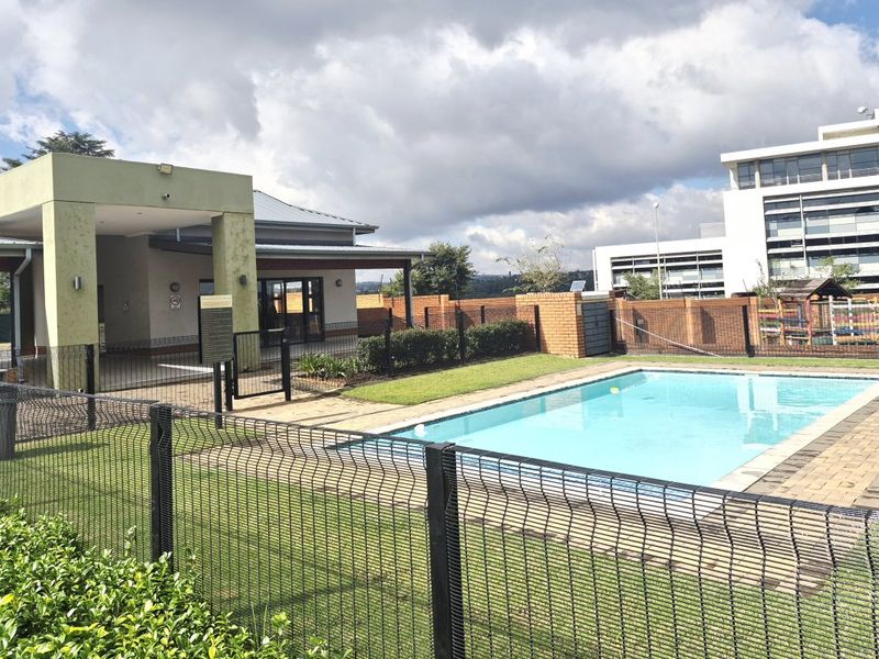 A well-maintained communal pool area featuring a paved surround, mesh fencing, and an adjacent covered patio space within an apartment complex.