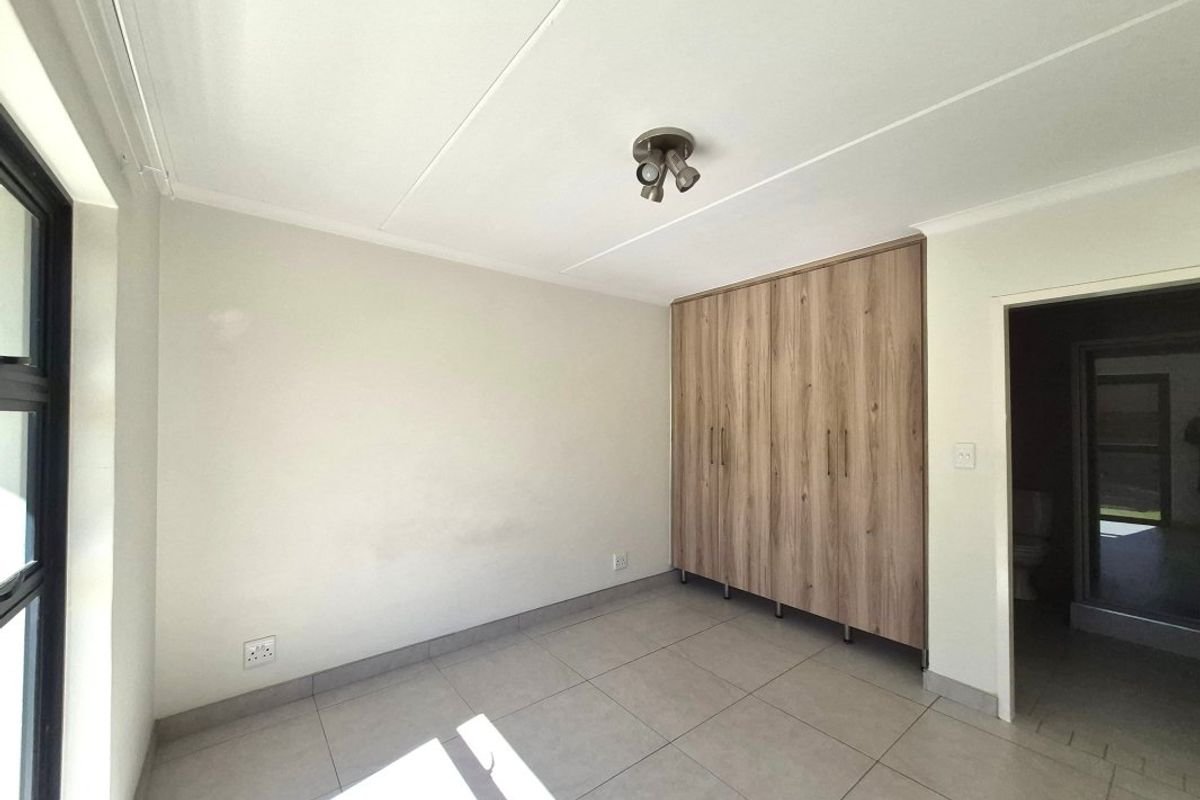 A bright, empty bedroom featuring neutral tiled flooring, modern wood-finish built-in cupboards, and a ceiling-mounted spotlight fixture.