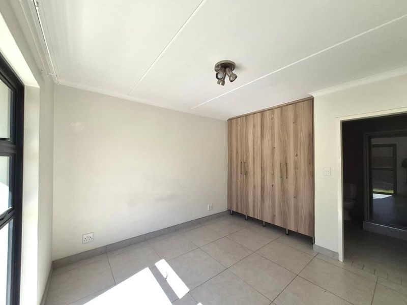 A bright, empty bedroom featuring neutral tiled flooring, modern wood-finish built-in cupboards, and a ceiling-mounted spotlight fixture.