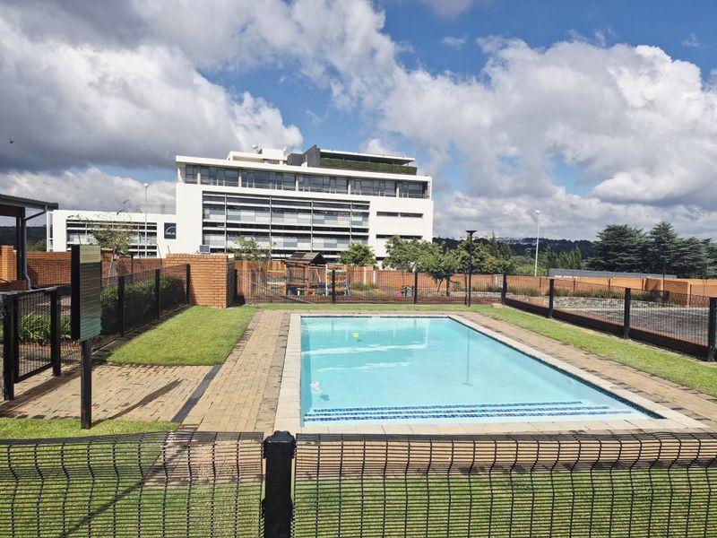 A well-maintained communal swimming pool area featuring paved surrounds, manicured lawn, and secure perimeter fencing within an apartment complex.