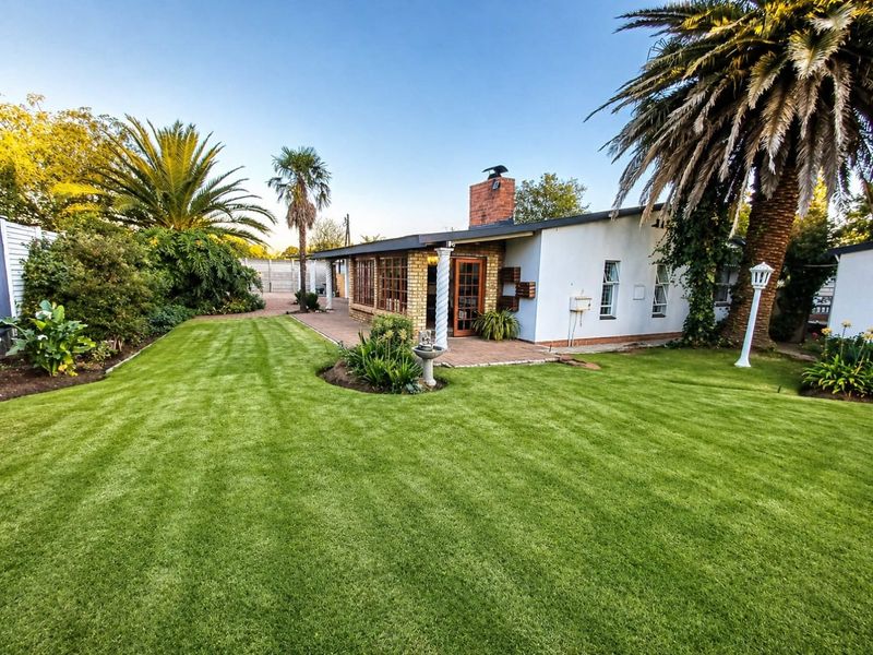 A well-maintained rear garden featuring a manicured lawn, paved patio area, and a brick chimney, surrounded by mature landscaping and palm trees.