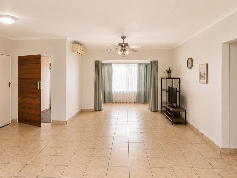 A spacious, neutrally painted living area featuring tiled flooring, a ceiling fan, and a wall-mounted air conditioning unit, offering a clean and versatile layout.