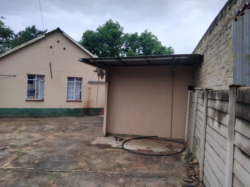 Rear exterior view showing a paved yard area, a lean-to structure, and the rear facade of the house with visible plaster cracking.