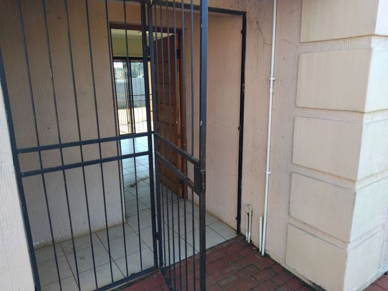 Entrance area featuring a metal security gate, tiled flooring, and brick paving, showing signs of wear on the exterior wall plaster.