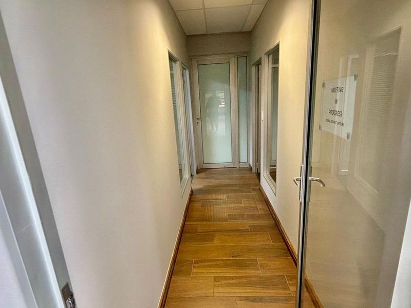 A clean, modern commercial hallway featuring wood-look tile flooring, glass partition doors, and a suspended ceiling, providing access to office spaces.