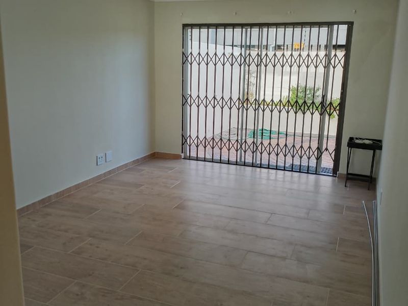 A bright, empty living room featuring modern wood-look floor tiles, recessed lighting, and a sliding glass door with a security gate leading to the patio.