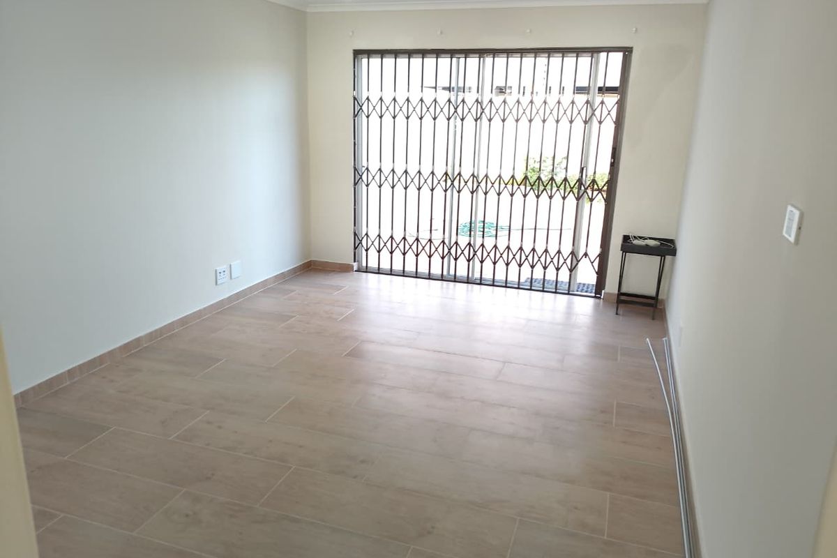 A bright, empty living room featuring modern tiled flooring, recessed downlights, and a sliding glass door with a security gate leading outdoors.