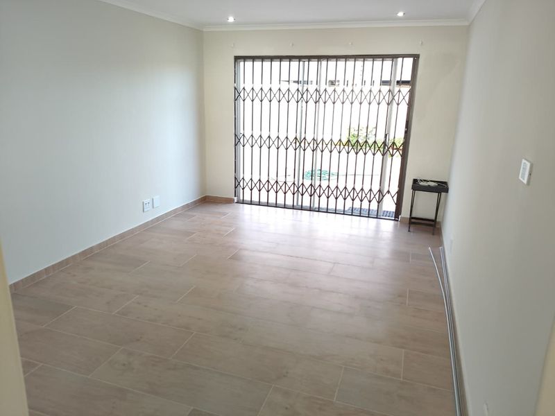 A bright, empty living room featuring modern tiled flooring, recessed downlights, and a sliding glass door with a security gate leading outdoors.