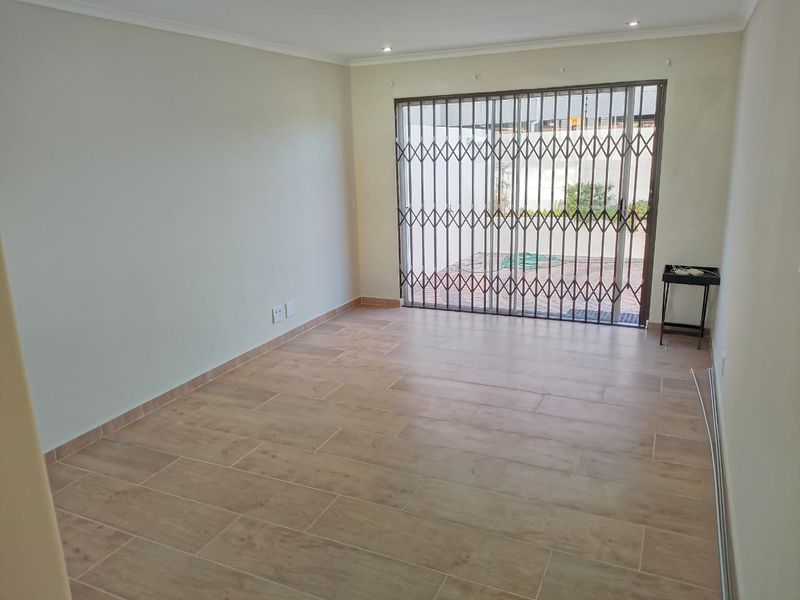 A bright, empty living room featuring modern wood-look floor tiles, recessed lighting, and a sliding glass door secured with a metal security gate.