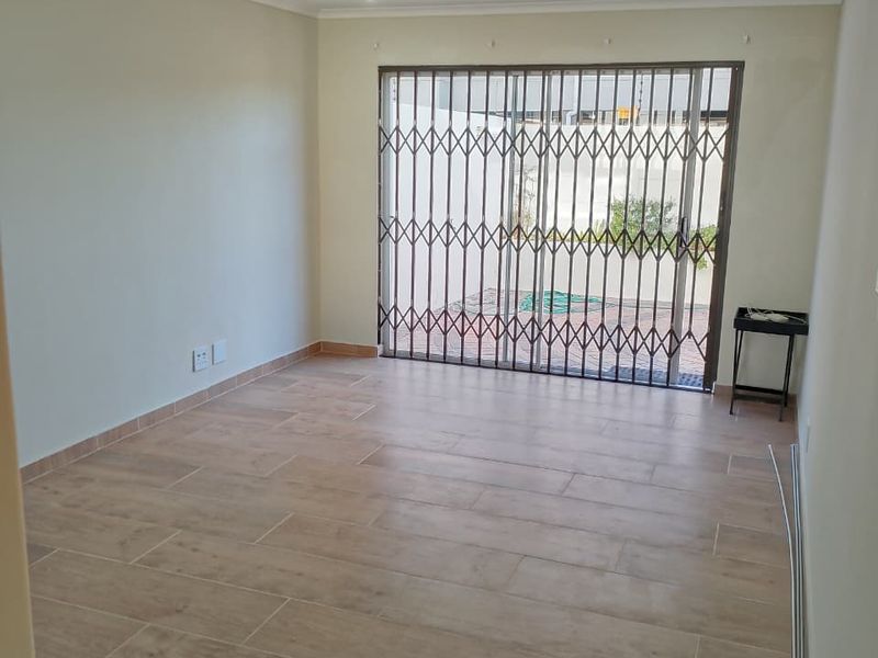 A bright, empty living room featuring modern wood-look tiled flooring, recessed downlights, and a secure sliding glass door leading to an outdoor area.