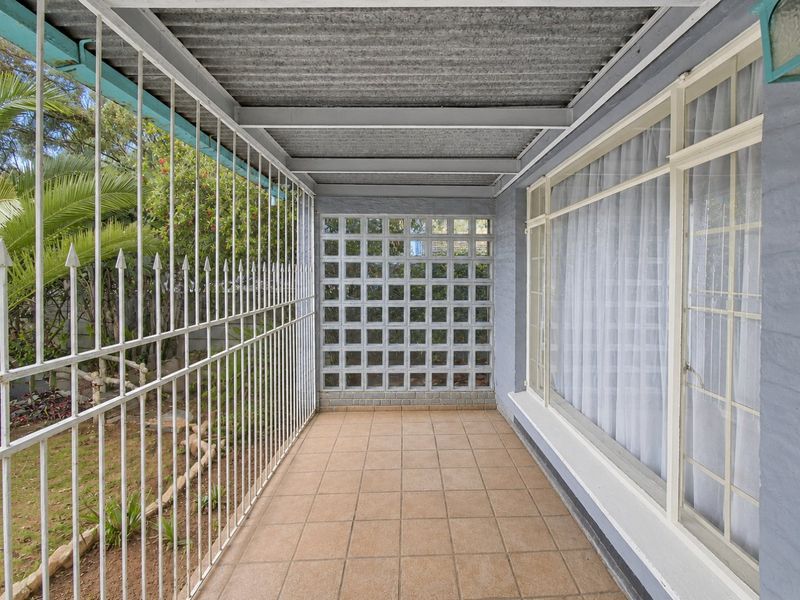 A covered patio area featuring tiled flooring, a corrugated metal roof, and security screening, overlooking a garden space.