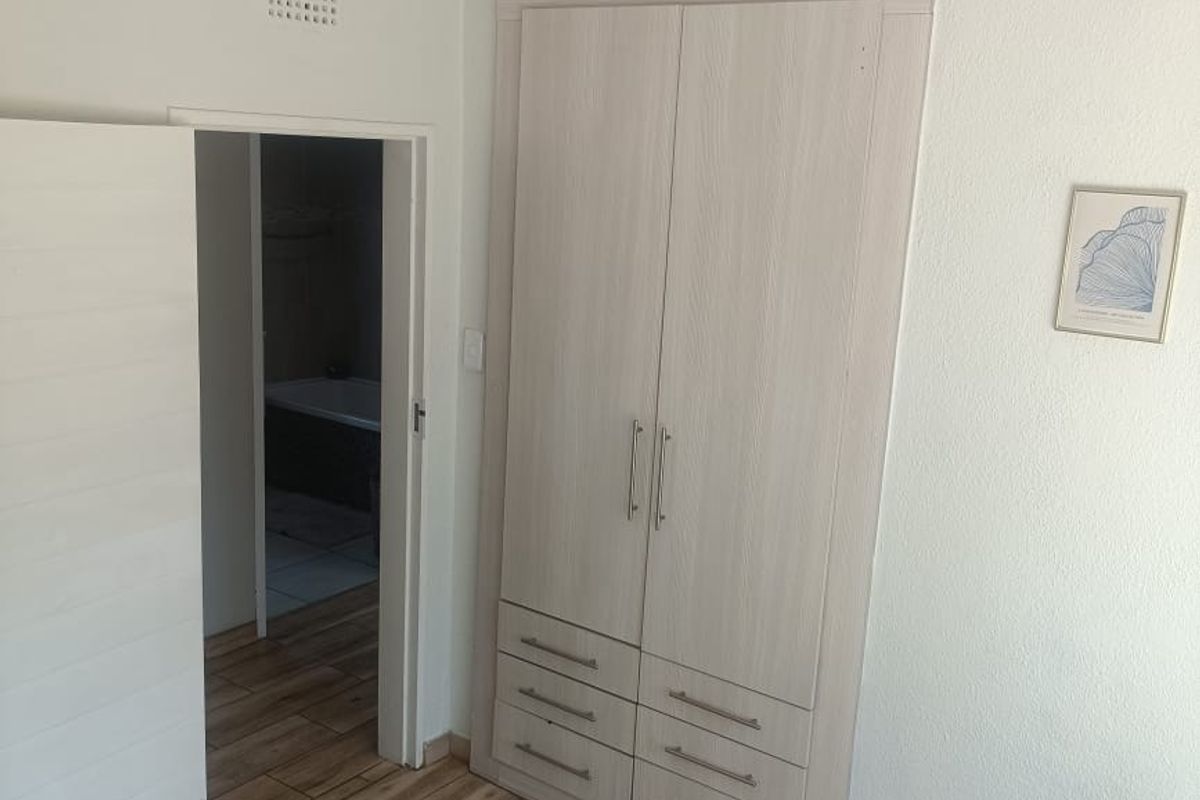 A clean bedroom featuring light wood-look laminate flooring, a modern built-in wardrobe, and recessed ceiling lighting, with an adjacent bathroom visible through the doorway.
