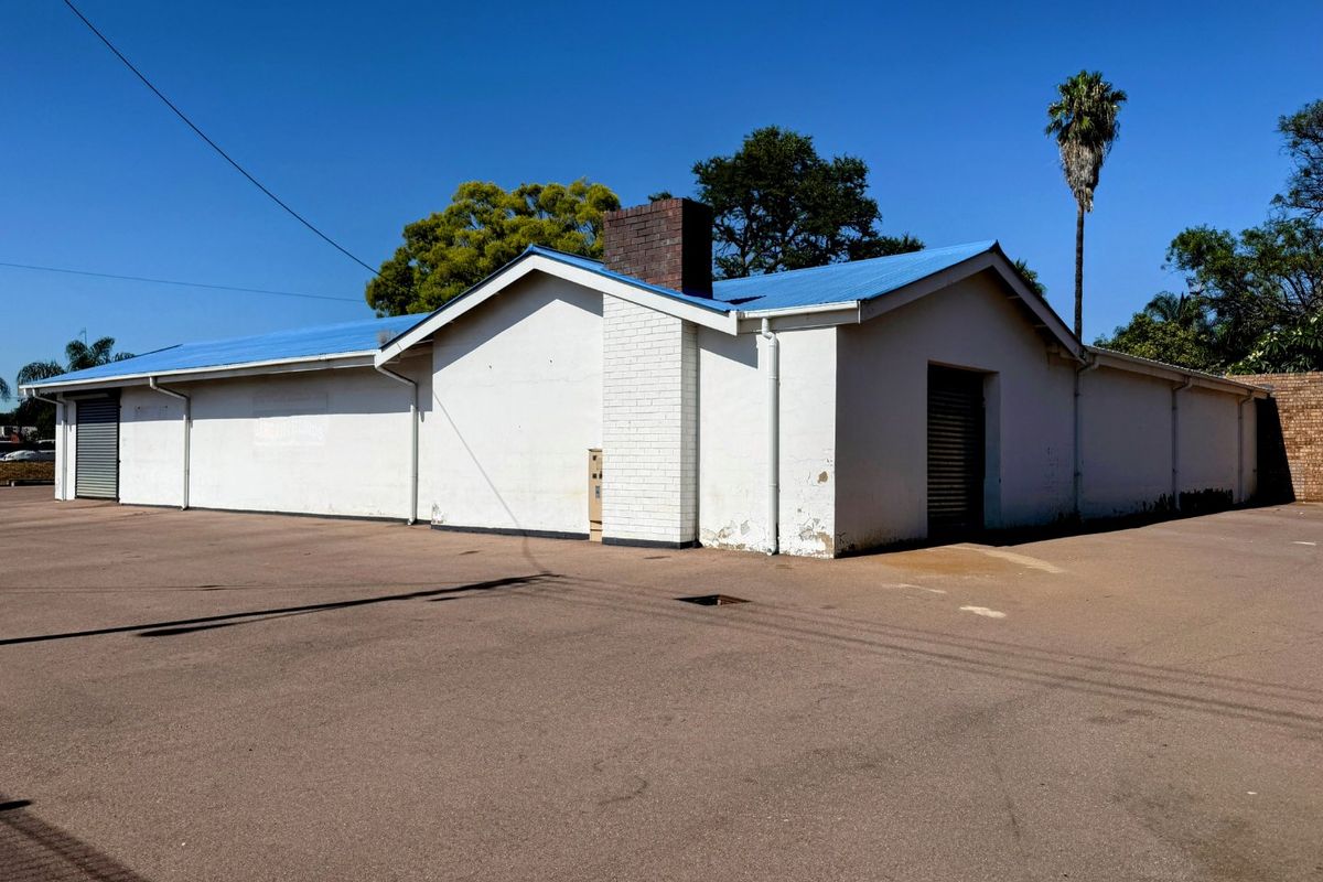 Rear view of a commercial building featuring a blue metal roof, brick chimney, and multiple roller shutter doors with an expansive asphalt parking area.