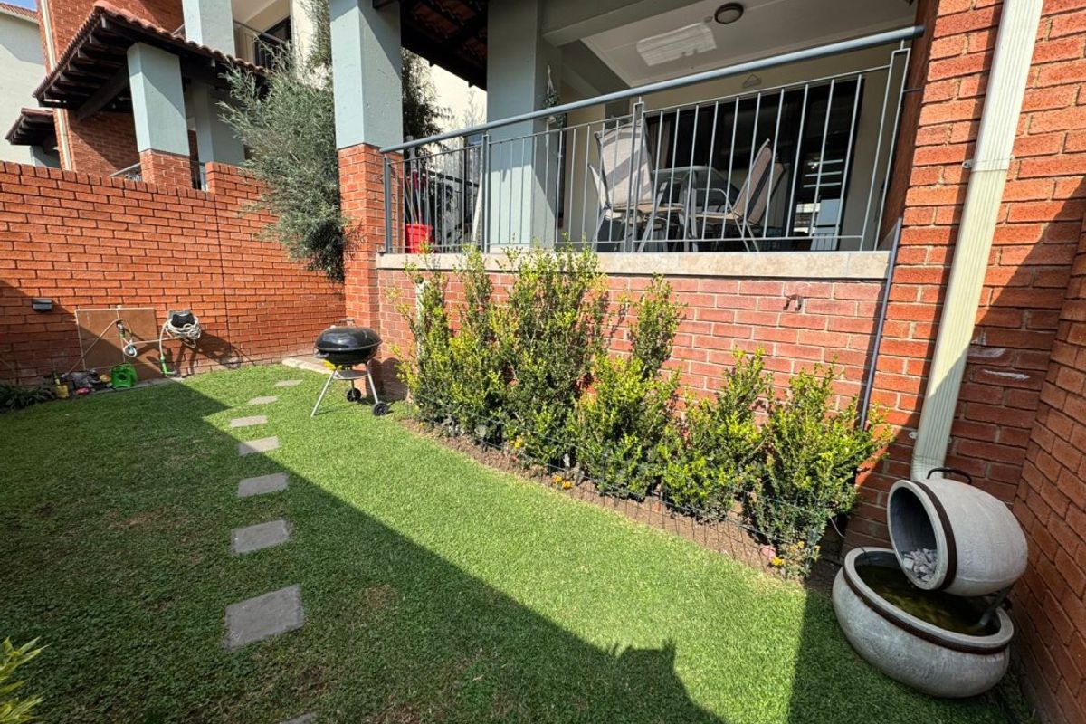 A private rear garden featuring a manicured lawn, stone water feature, and a raised patio area with metal balustrades and brick walling.