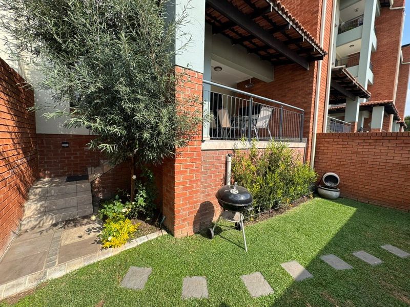 A private rear garden area featuring a paved patio with metal balustrade, brick exterior walls, and a well-maintained lawn with stone stepping stones.