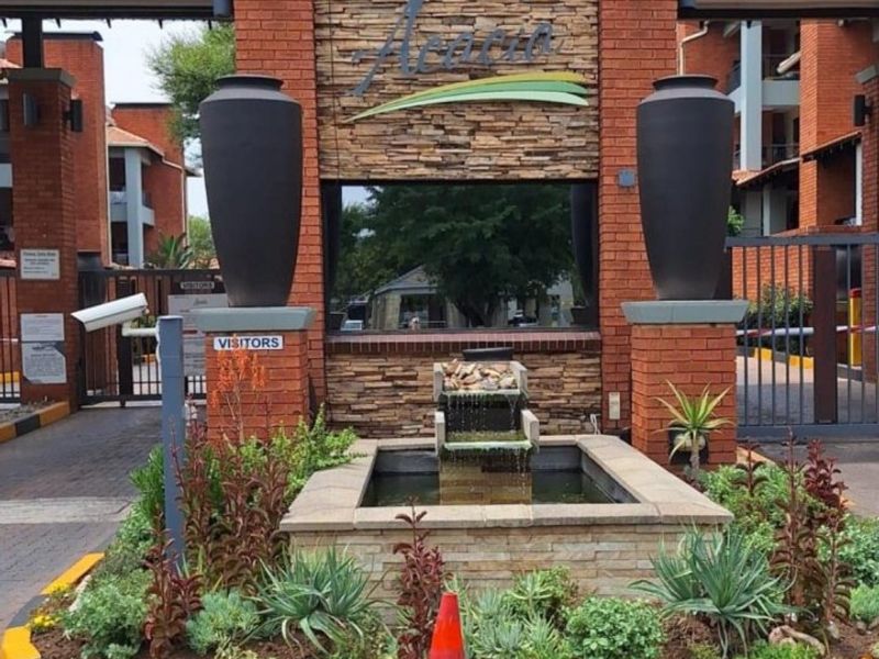 The entrance to the Acacia complex features a brick and stone facade, a decorative water feature, and secure gated access.