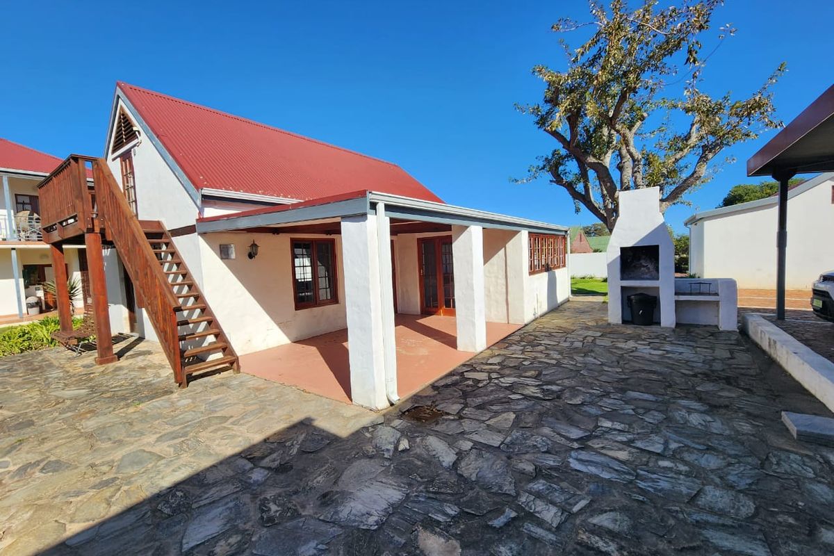 Rear exterior view featuring a covered patio, built-in braai, stone paved courtyard, and an external wooden staircase leading to the upper level.