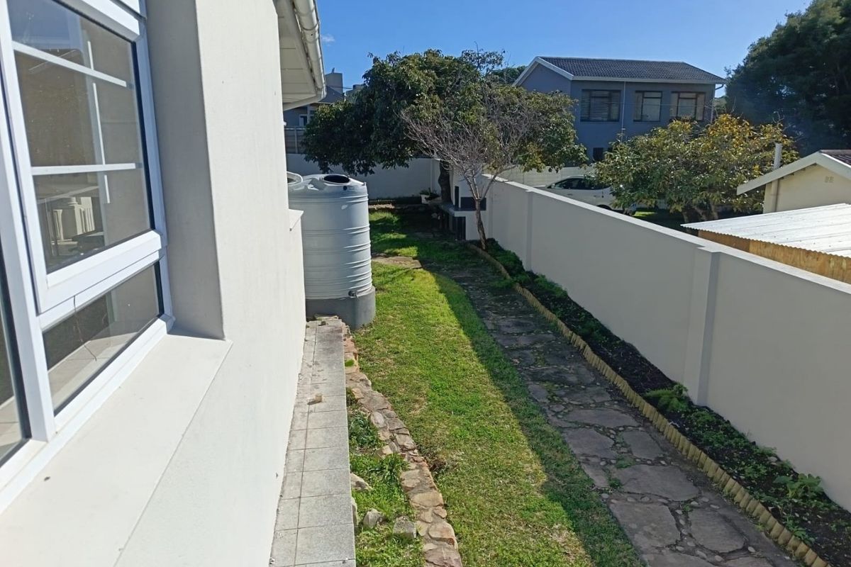 A side garden view featuring a water storage tank, stone pathway, and a boundary wall with a built-in braai area.