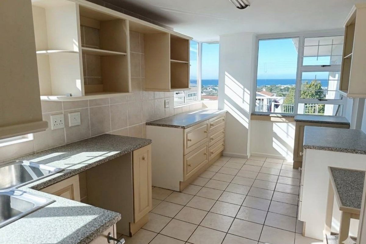 A bright, empty kitchen featuring tiled flooring, ample built-in cabinetry, laminate countertops, and large windows offering scenic coastal views.
