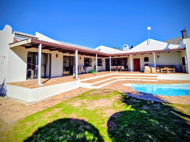 Spacious rear exterior featuring a covered patio, brick-paved entertainment area, and a swimming pool set against a white-walled house structure.