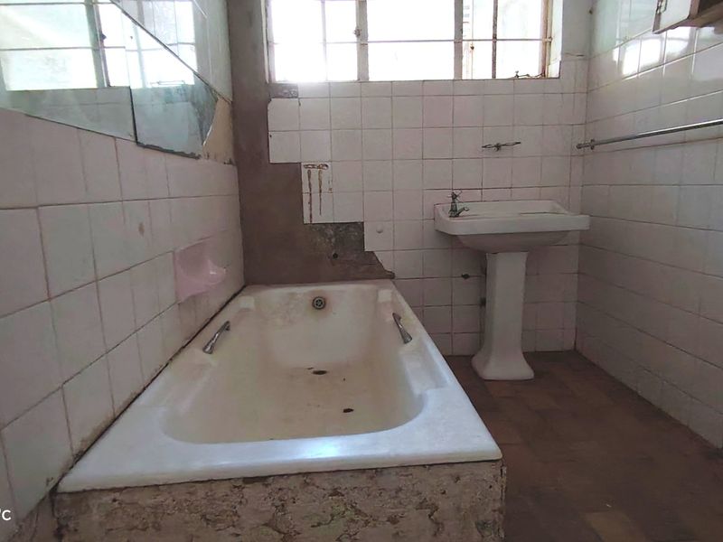 A dated bathroom requiring significant renovation, featuring a pedestal basin and built-in bathtub with missing wall tiles and damaged finishes.