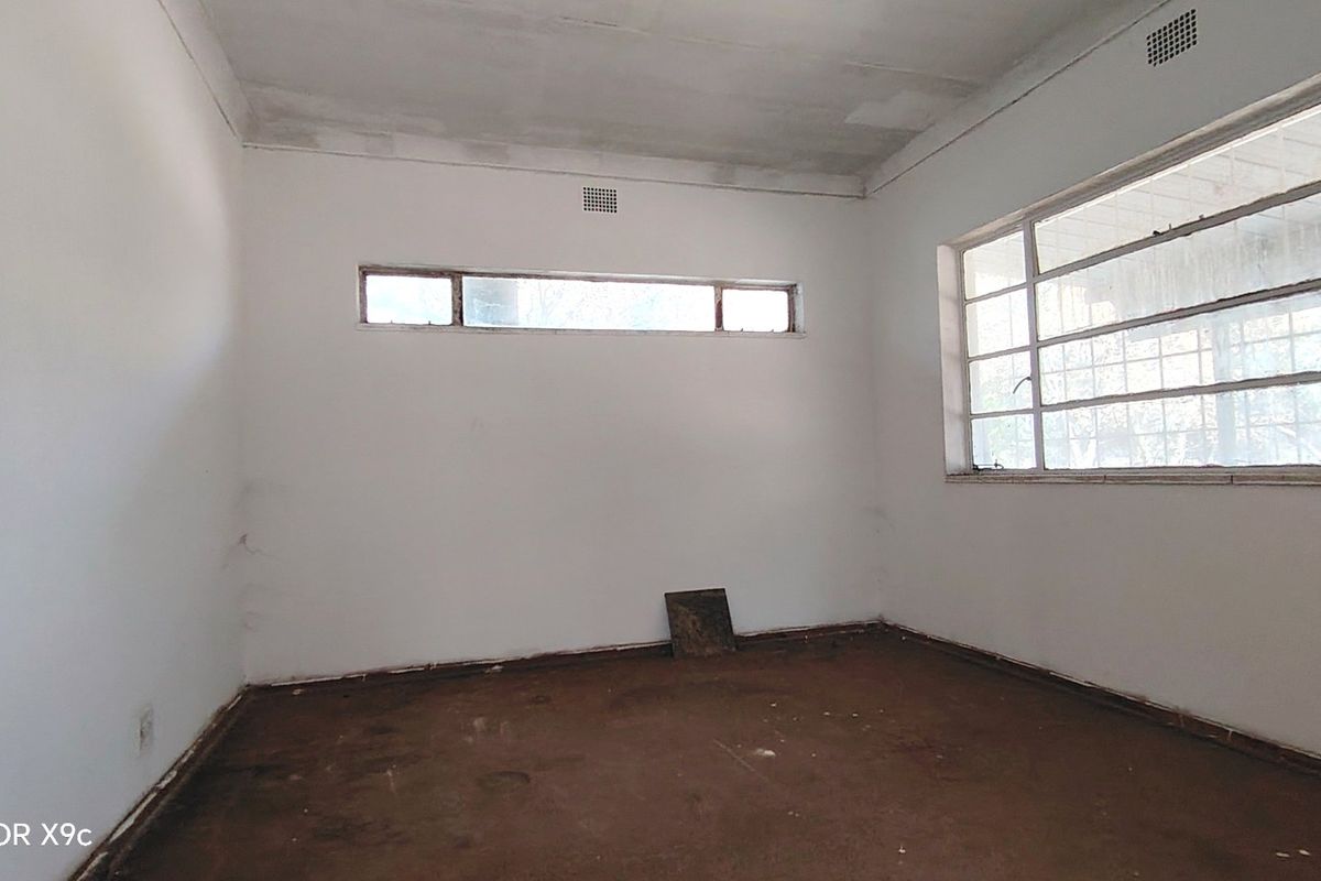 An empty, unrenovated bedroom featuring steel-framed windows and ventilation grilles, requiring significant cosmetic updates and flooring replacement.