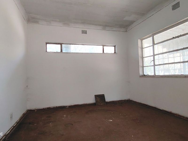 An empty, unrenovated bedroom featuring steel-framed windows and ventilation grilles, requiring significant cosmetic updates and flooring replacement.
