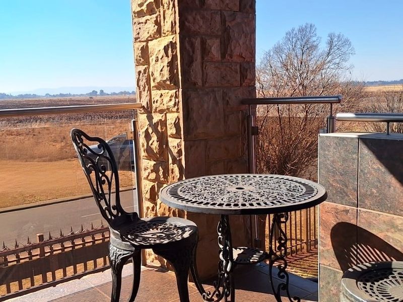 A covered balcony featuring stone pillar accents, tiled finishes, and a modern stainless steel and glass balustrade overlooking the surrounding landscape.