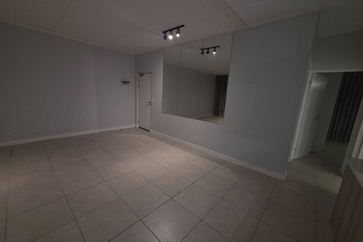 An empty, neutrally painted living area featuring large format floor tiles, a wall-mounted mirror, and modern track lighting.
