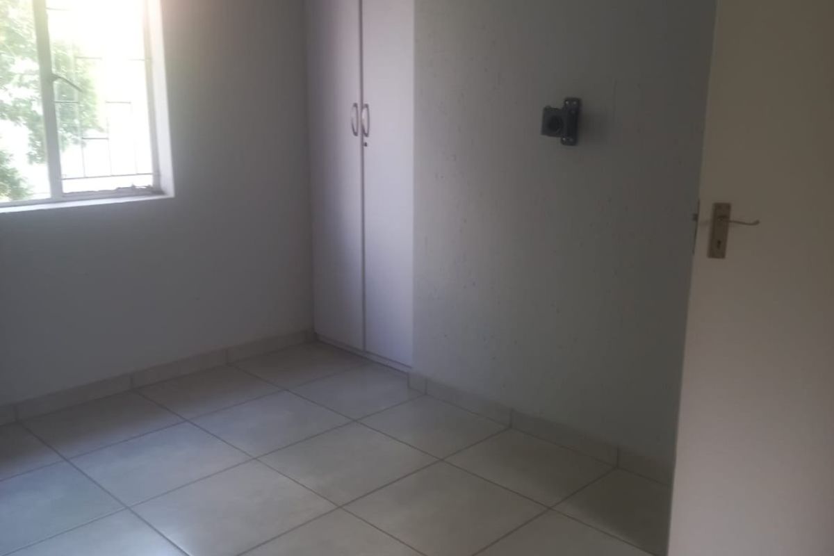 A bright, empty bedroom featuring neutral tiled flooring, a built-in wardrobe, and a window fitted with security bars.
