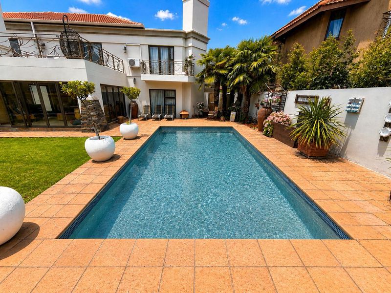 A well-maintained outdoor pool area featuring a tiled deck, landscaped garden, and a multi-level home exterior with a balcony and wrought iron railings.