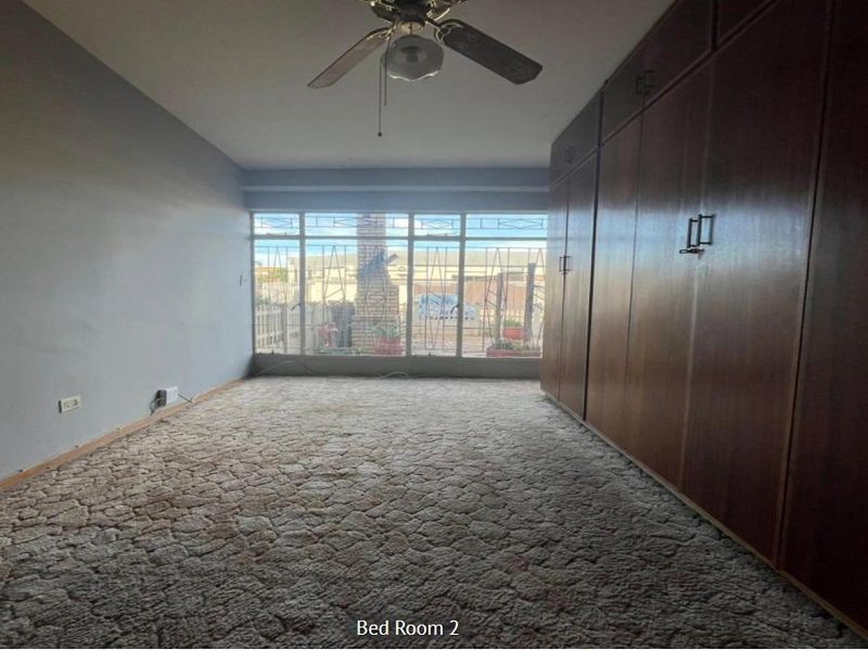 Spacious bedroom featuring extensive built-in wooden cupboards, a ceiling fan, and large windows with security bars, offering a blank canvas for renovation.