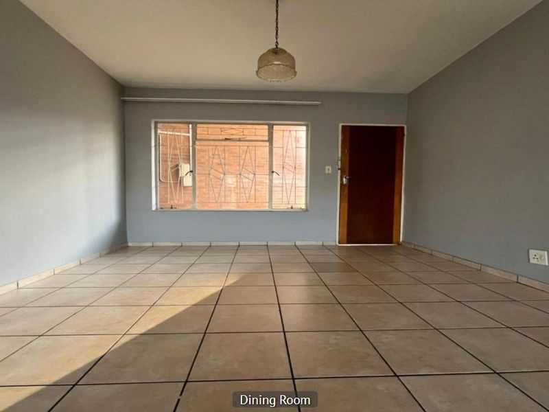An empty dining room featuring neutral grey walls, tiled flooring, a central pendant light, and a window fitted with security bars.