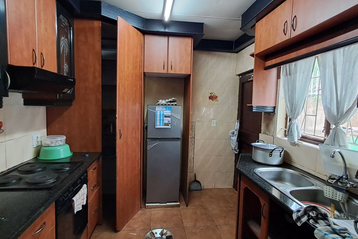 A dated kitchen featuring wood-tone cabinetry, dark countertops, an electric hob, and a double bowl sink with tiled splashbacks.