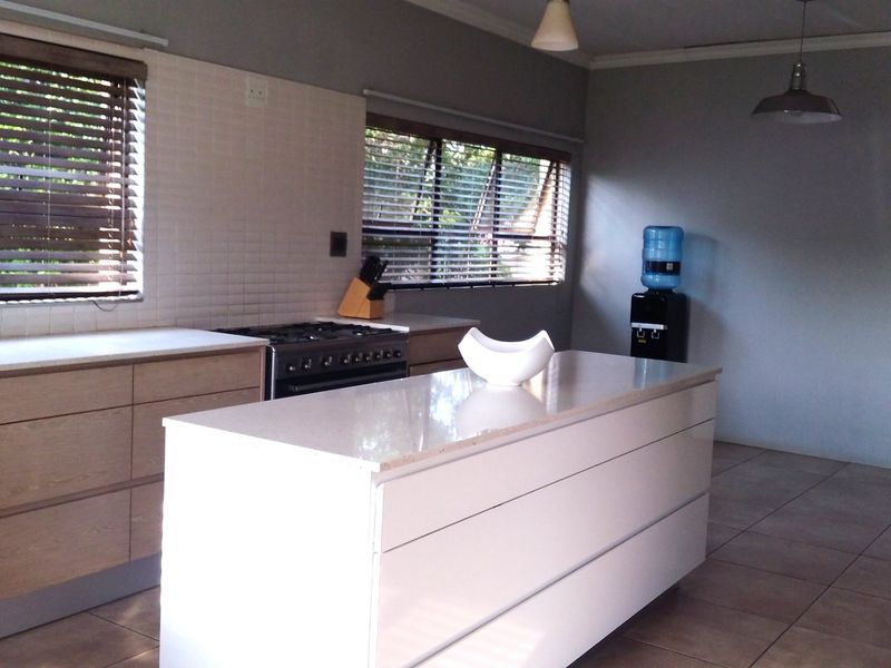 Modern kitchen featuring a central island with stone countertops, gas stove, light wood cabinetry, and tiled backsplash with natural light from venetian blinds.