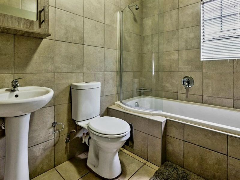 A functional bathroom featuring a pedestal basin, toilet, and a bathtub with a glass shower screen, finished with neutral wall and floor tiles.