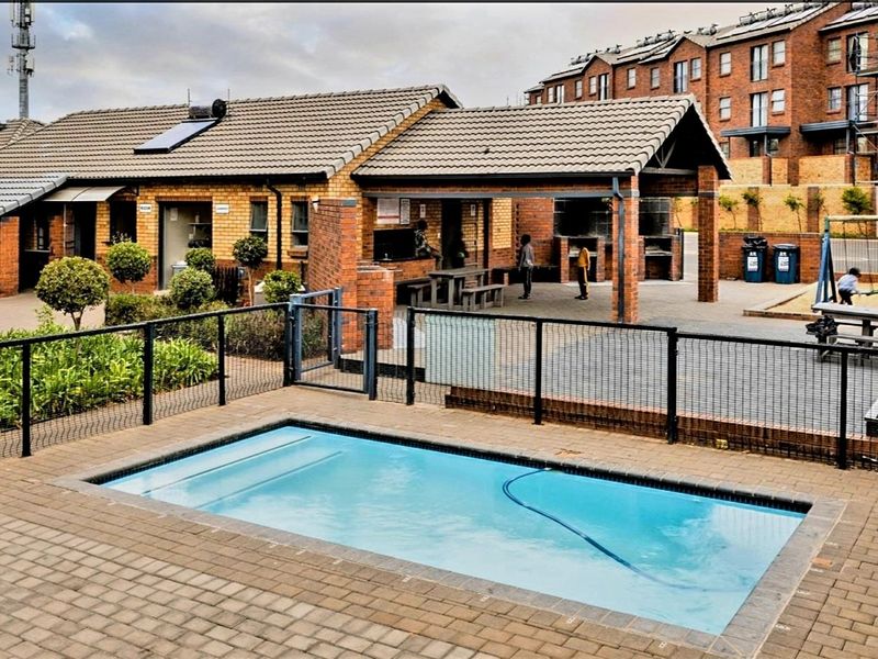 Communal pool area featuring brick paving, a covered patio with built-in braai facilities, and secure metal fencing within an apartment complex.